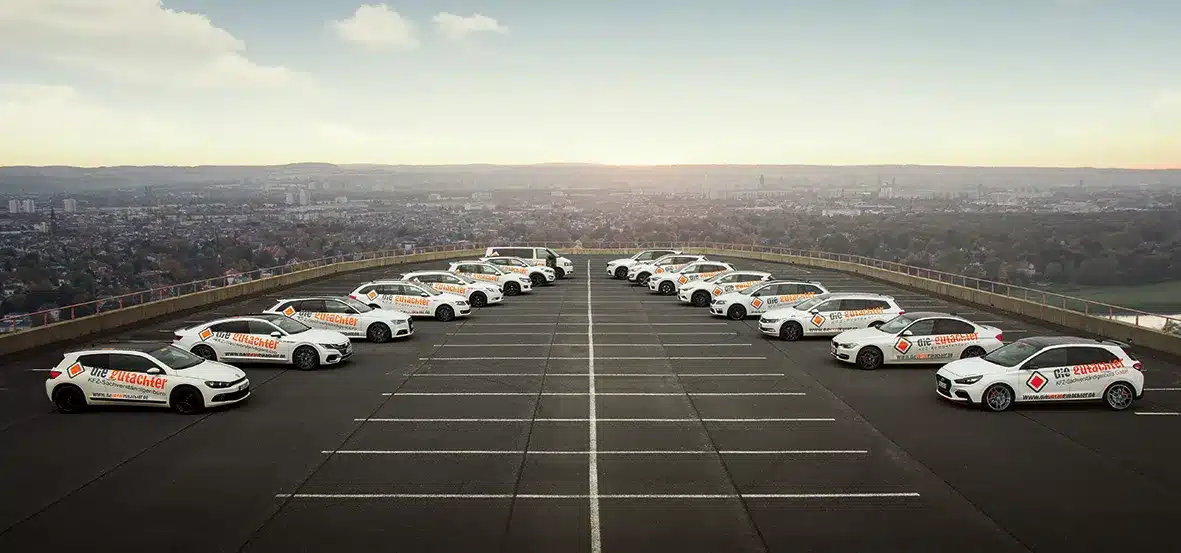 A fleet of white cars with "GoStudent" branding is parked in a semi-circle on an empty rooftop parking lot at sunset, ideal for a kfz sachverständiger or unfallgutachter Dresden to assess vehicles against the stunning cityscape backdrop.