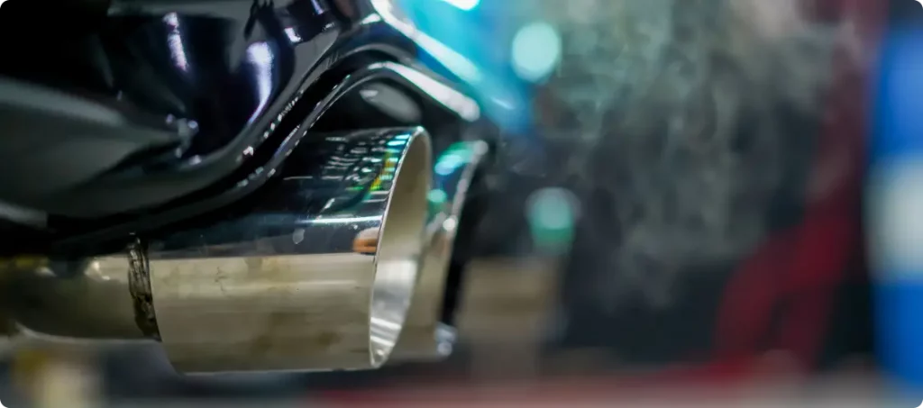 Nahaufnahme des glänzenden Metallauspuffs eines Autos mit sichtbaren Abgasen vor einem unscharfen Hintergrund.