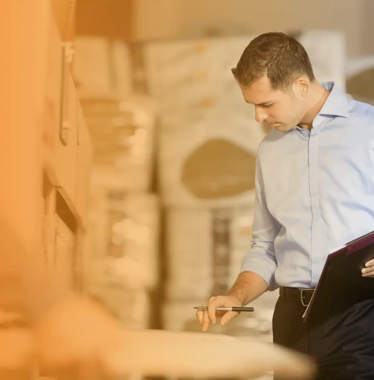 A kfz sachverständiger in a light blue shirt holds a clipboard and pen while inspecting boxes in a warehouse filled with stacked packages.