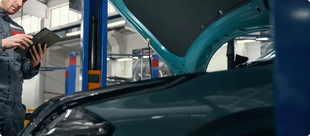 Ein Mechaniker in einer Werkstatt steht neben einem Auto mit geöffneter Motorhaube und hält ein Tablet in der Hand, während er das Fahrzeug inspiziert oder eine Diagnose stellt. Das Bild konzentriert sich auf das Auto und die untere Hälfte des Mechanikers.