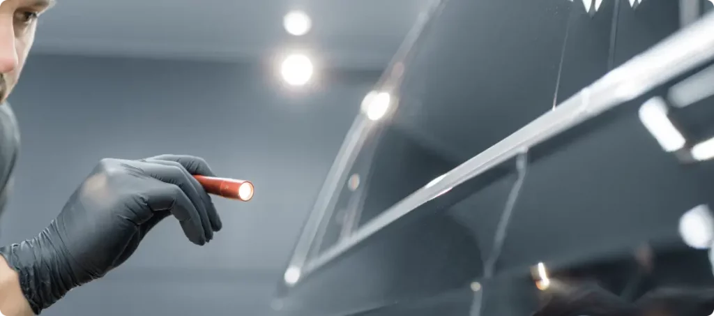 Eine Person, die einen schwarzen Handschuh trägt, prüft die Oberfläche eines glänzenden schwarzen Autos mit einer kleinen Taschenlampe auf Unvollkommenheiten oder Kratzer.