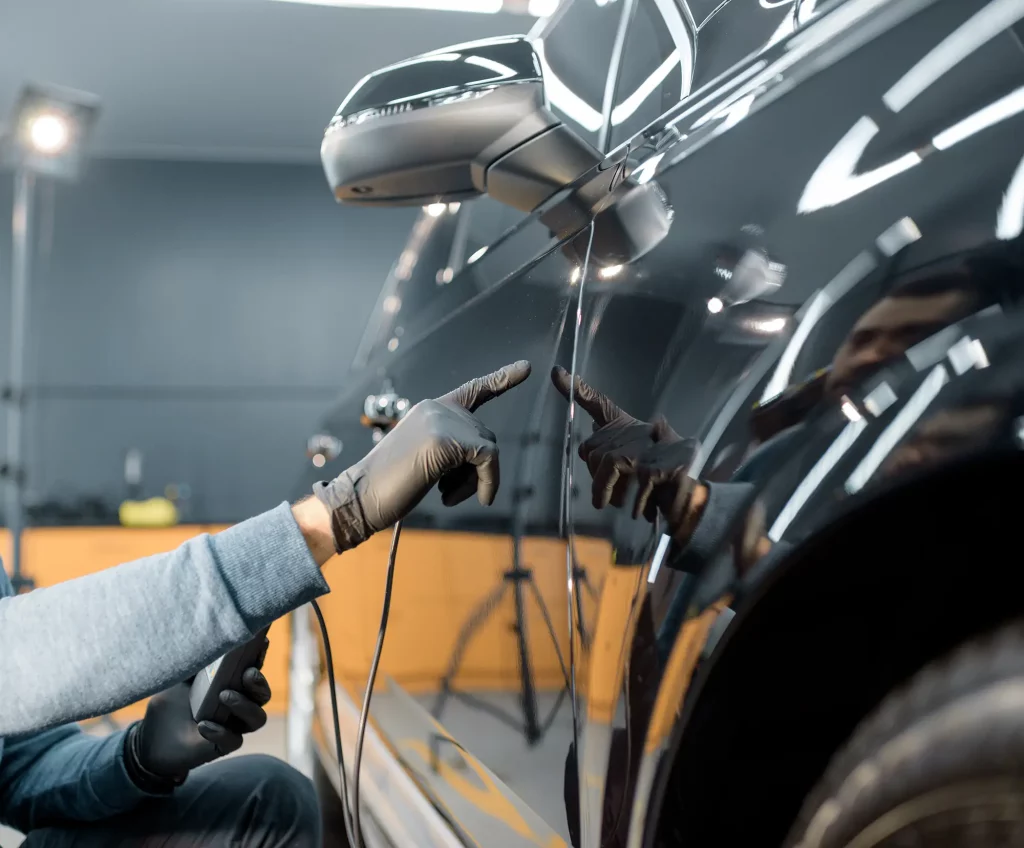 Eine Person mit schwarzen Handschuhen zeigt auf einen Kratzer an der Seite eines glänzenden schwarzen Autos, das in einer gut beleuchteten Garage aus der Nähe begutachtet wird. Die Szene wirkt wie ein Moment im Umfeld einer Auto-Hauptuntersuchung oder Abgasuntersuchung in Dresden, bei der Fahrzeugzustand und sicherheitsrelevante Details geprüft werden.