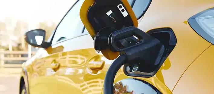 A yellow electric car is being charged, with a charging cable plugged into its port on the rear side of the vehicle. In the slightly blurred background, a kfz sachverständiger prepares a schadensgutachten.