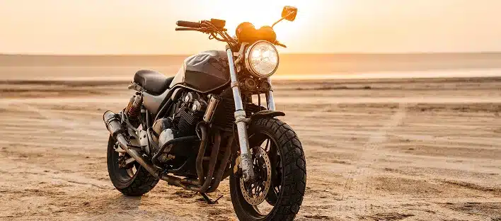 A black motorcycle is parked on sandy terrain at sunset, with the sun low on the horizon. In the open landscape, a kfz gutachter dresden could expertly assess this scenic scene for any passionate rider.