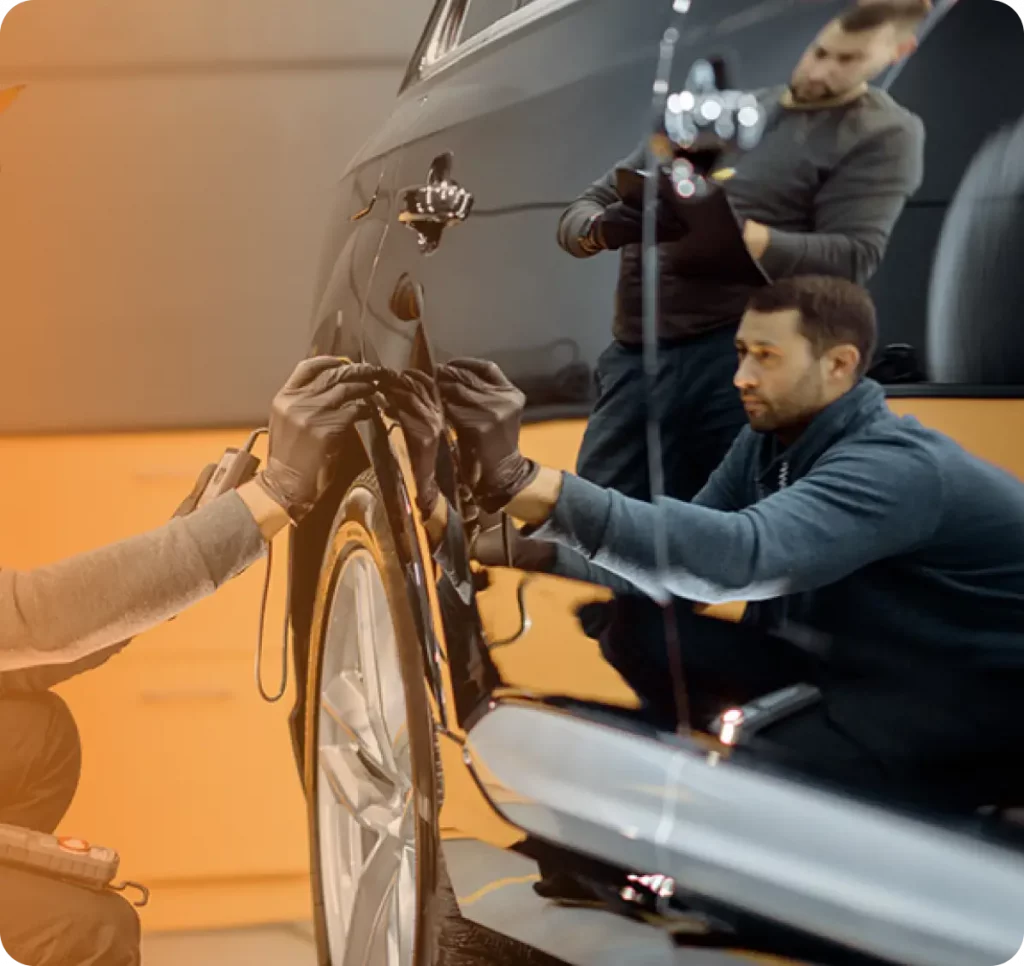 Zwei Männer mit Handschuhen arbeiten an einem glänzenden schwarzen Auto in einem Innenraum, wobei sie sich auf der Oberfläche des Autos spiegeln.