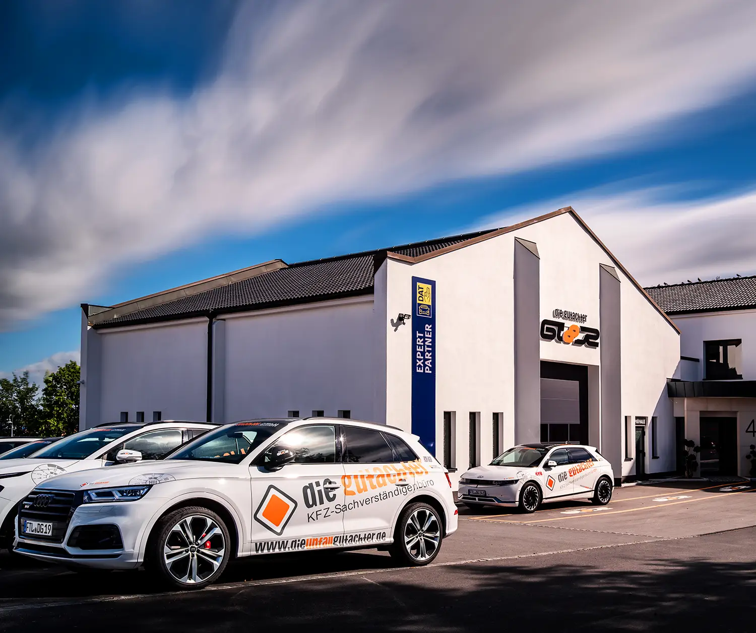 Modernes weißes Gebäude mit „GT8R“-Schild, davor parken mehrere weiße Autos mit Markenlogo, darüber dramatische Wolken. Die Autos und das Gebäude sind mit Logos und deutschem Text versehen, die auf die Dienstleistungen von KFZ Gutachter Dresden hinweisen.