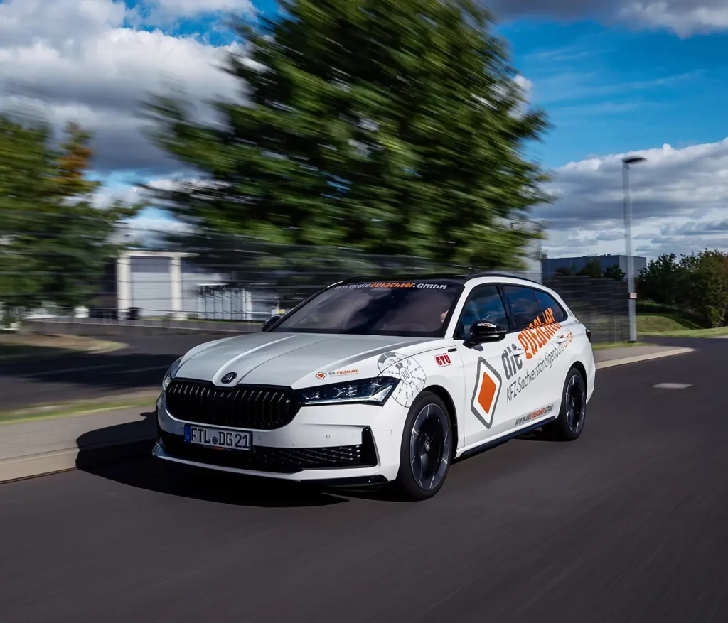 Ein weißer Skoda mit dem Schriftzug "die Gutachter" auf einer Straße, die von Grünflächen und Gebäuden umgeben ist, unter einem teilweise bewölkten Himmel. Das Bild fängt das Auto in Bewegung ein.