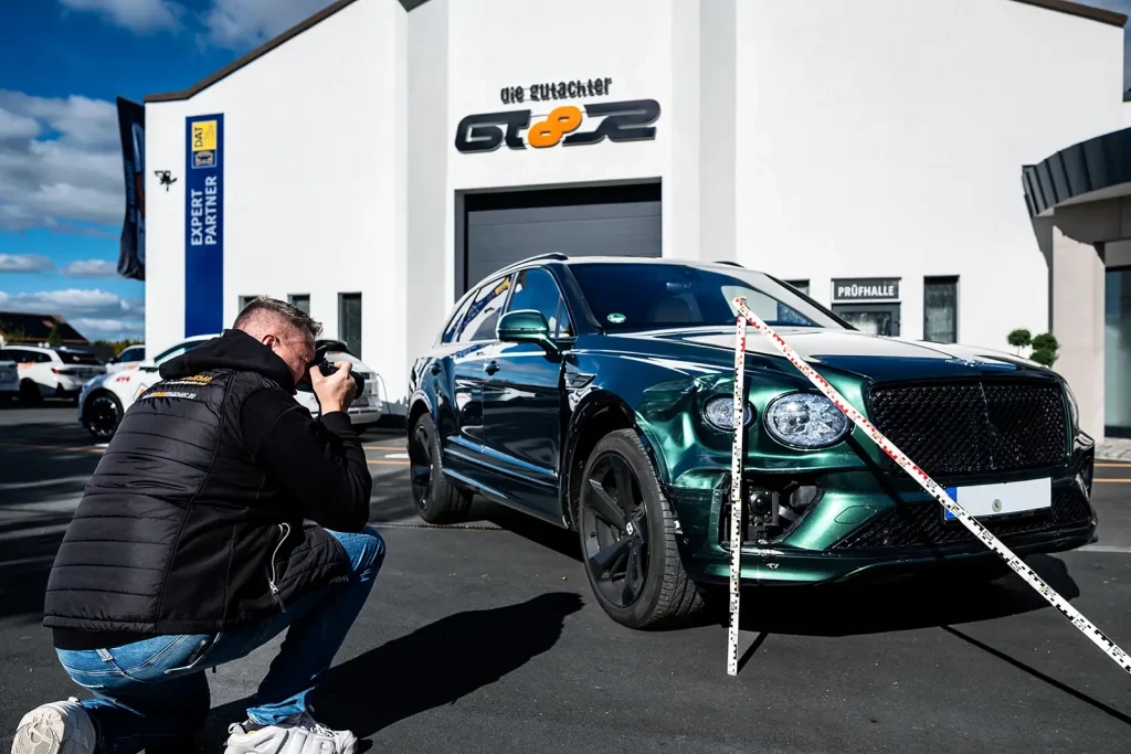 Ein Gutachter kniet nieder, um ein Foto von einem glänzenden grünen Bentley SUV zu machen, der vor einem weißen Gebäude mit "GT8R"-Schriftzug unter blauem Himmel geparkt ist. Es werden die Schäden dokumentiert.