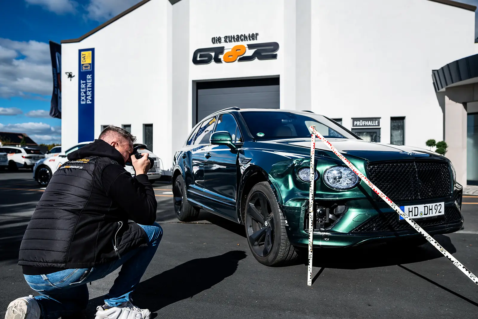 Ein Gutachter kniet nieder, um ein Foto von einem glänzenden grünen Bentley SUV zu machen, der vor einem weißen Gebäude mit "GT8R"-Schriftzug unter blauem Himmel geparkt ist. Der Schriftzug KFZ Gutachter Dresden ist in der Nähe des gestreiften Absperrbandes vor dem Auto zu sehen.