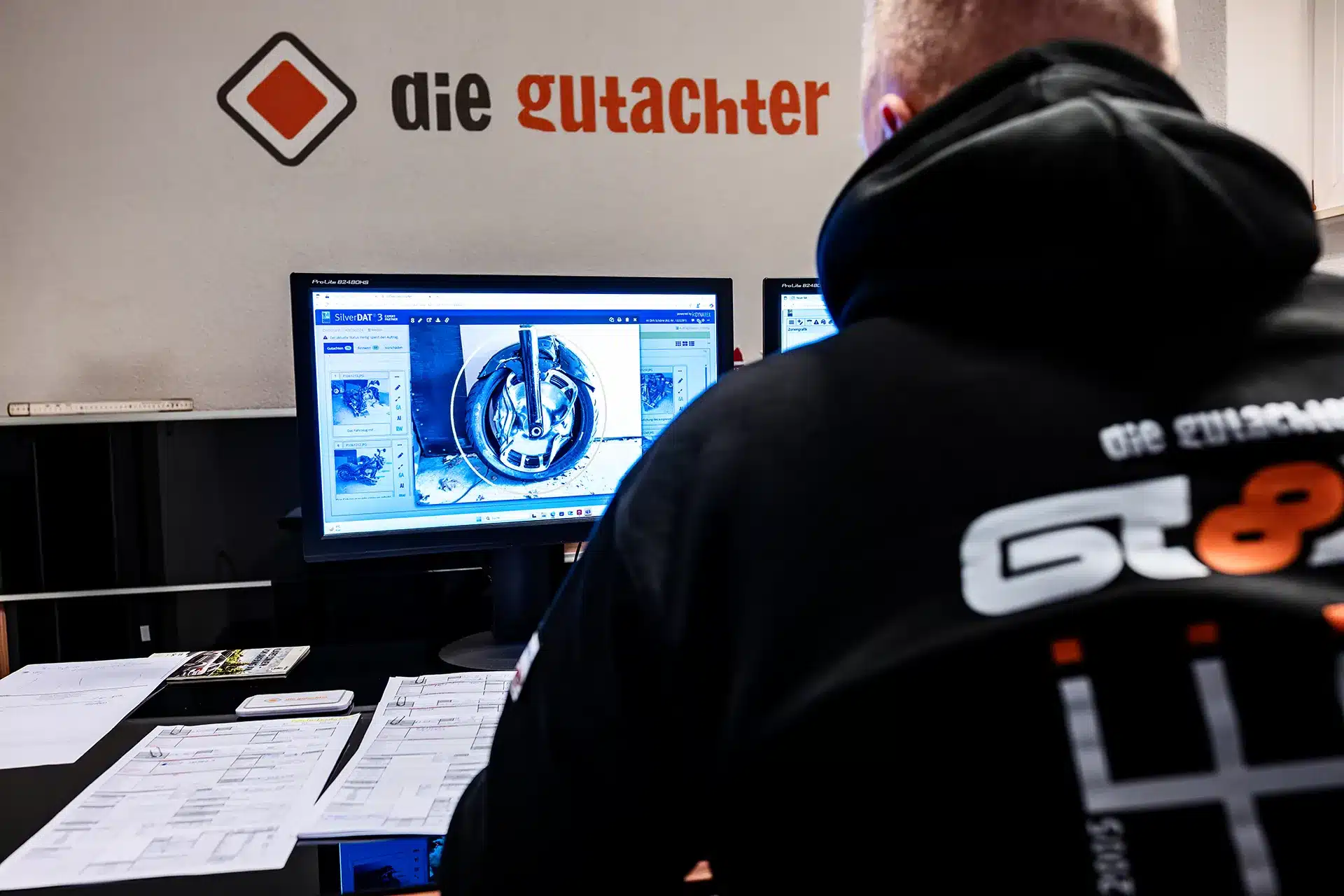 Eine Person in einem schwarzen Kapuzenpulli sitzt an einem Schreibtisch mit Papieren und blickt auf zwei Monitore, auf denen ein Autorad in Großaufnahme zu sehen ist. An der Wand sind das Logo und der Text "die gutachter.