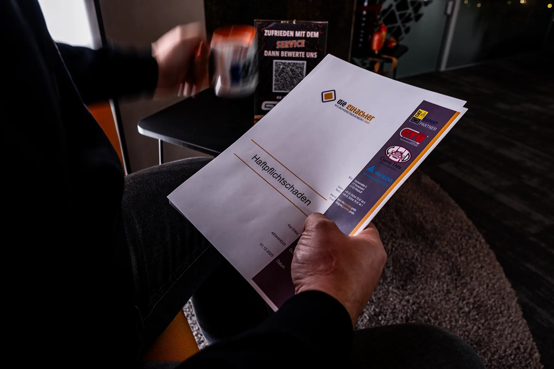 Eine Person hält Dokumente mit der Aufschrift "Haftpflichtschaden" in der Hand und sitzt in einem Haus. Im Hintergrund sind auf einem Tisch ein QR-Code und ein Schild mit einer Servicebewertung zu sehen. Die Szene scheint in einem Büro oder Wartebereich zu spielen.