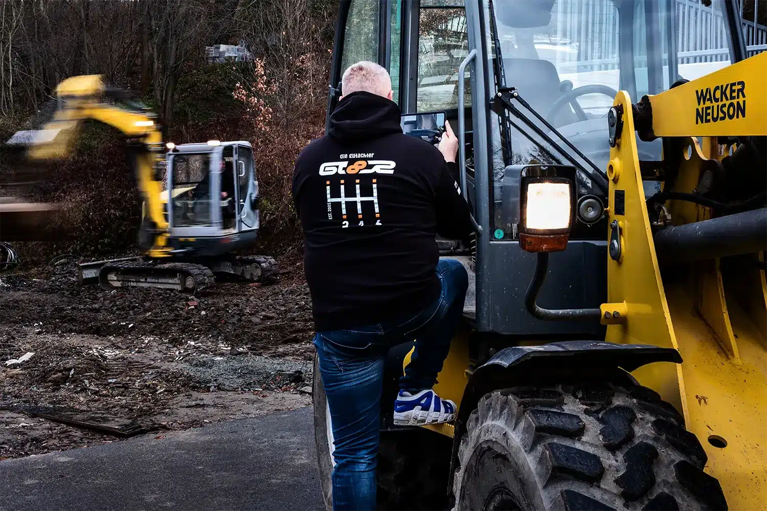 Ein Mann mit schwarzem Kapuzenpulli klettert auf ein gelbes Baufahrzeug auf einer Baustelle, während im Hintergrund ein anderer Bagger inmitten von Erde und Bäumen arbeitet.