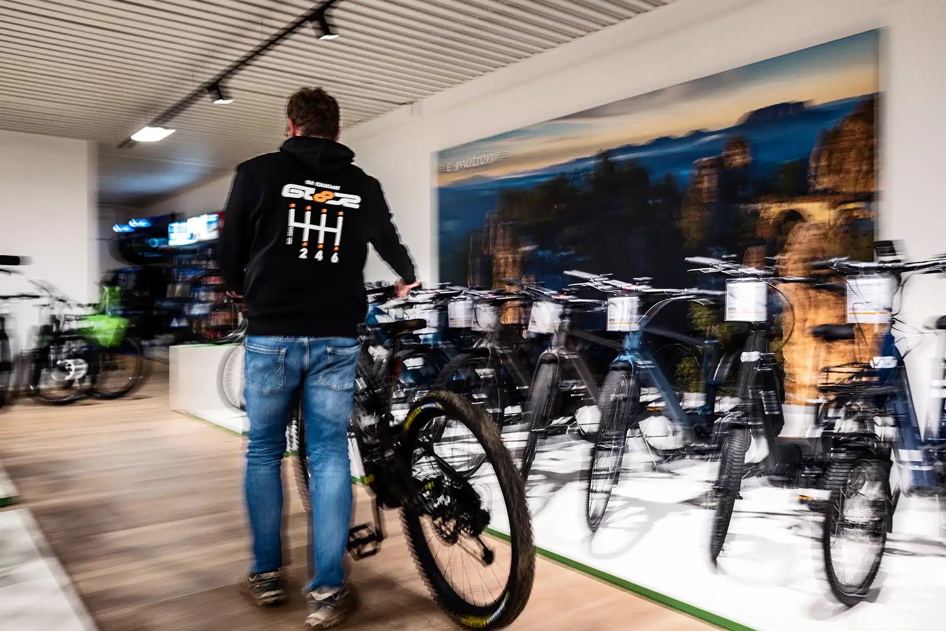 Ein Mann in Jeans und schwarzem Kapuzenpulli fährt mit einem Fahrrad durch ein Fahrradgeschäft, vorbei an Reihen von ausgestellten Fahrrädern. Die Szene ist leicht unscharf und suggeriert Bewegung.
