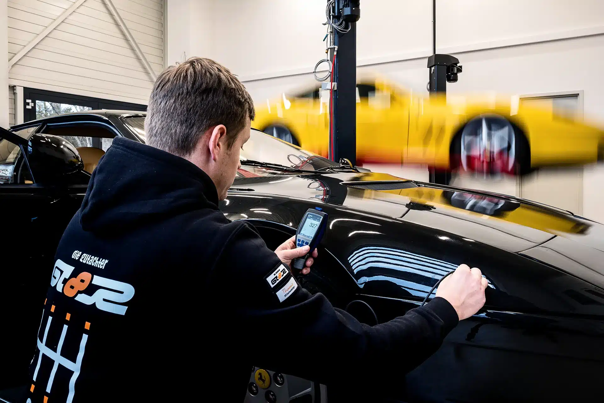 Ein Mann inspiziert mit einem Handgerät die Oberfläche eines schwarzen Autos in einer Garage, während im Hintergrund ein gelber Sportwagen angehoben wird.
