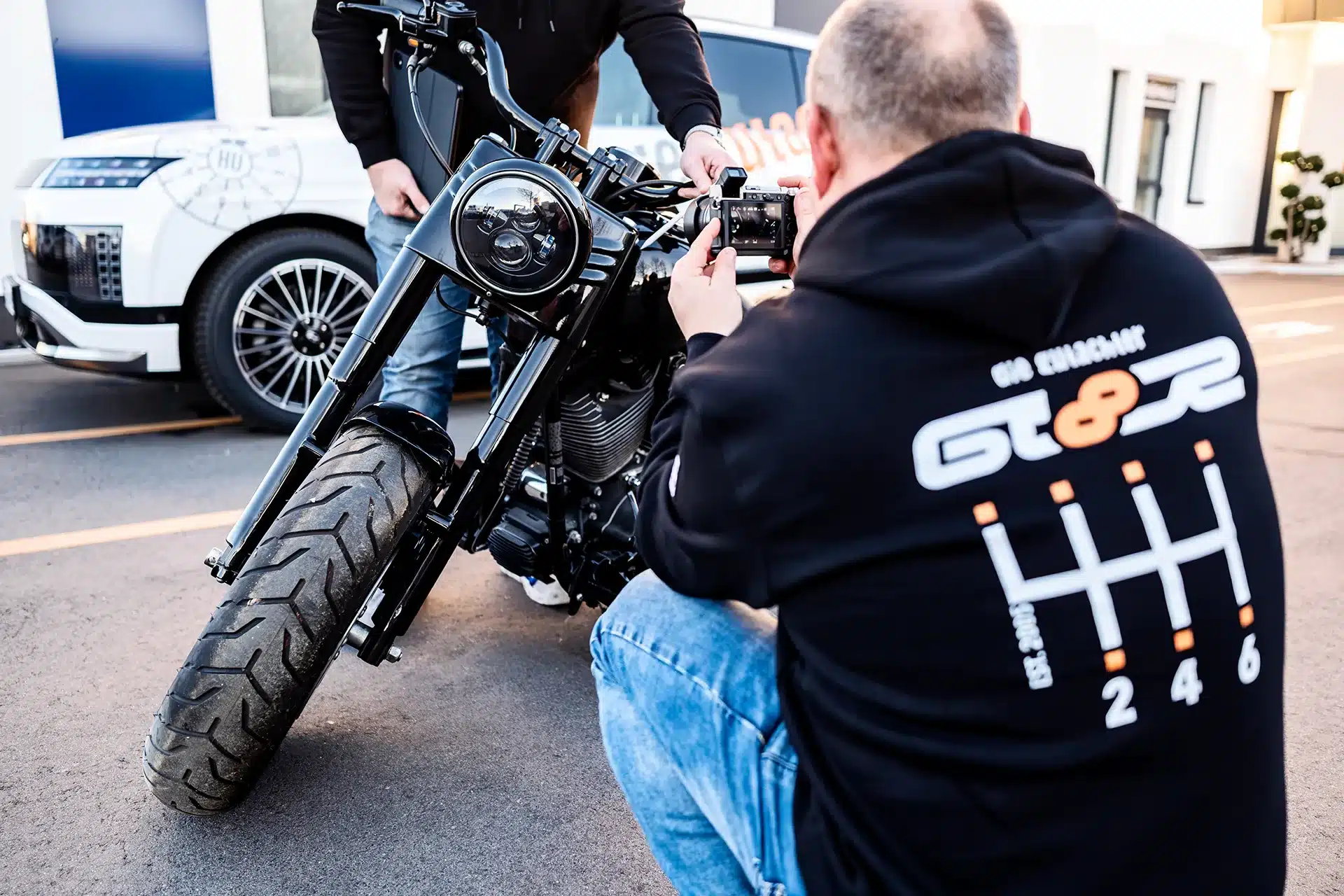 Eine Person in einem schwarzen Kapuzenpulli hockt sich hin und fotografiert ein schwarzes Motorrad, das auf der Straße geparkt ist, während eine andere Person neben dem Motorrad steht. Im Hintergrund ist ein weißes Auto mit Grafik zu sehen.