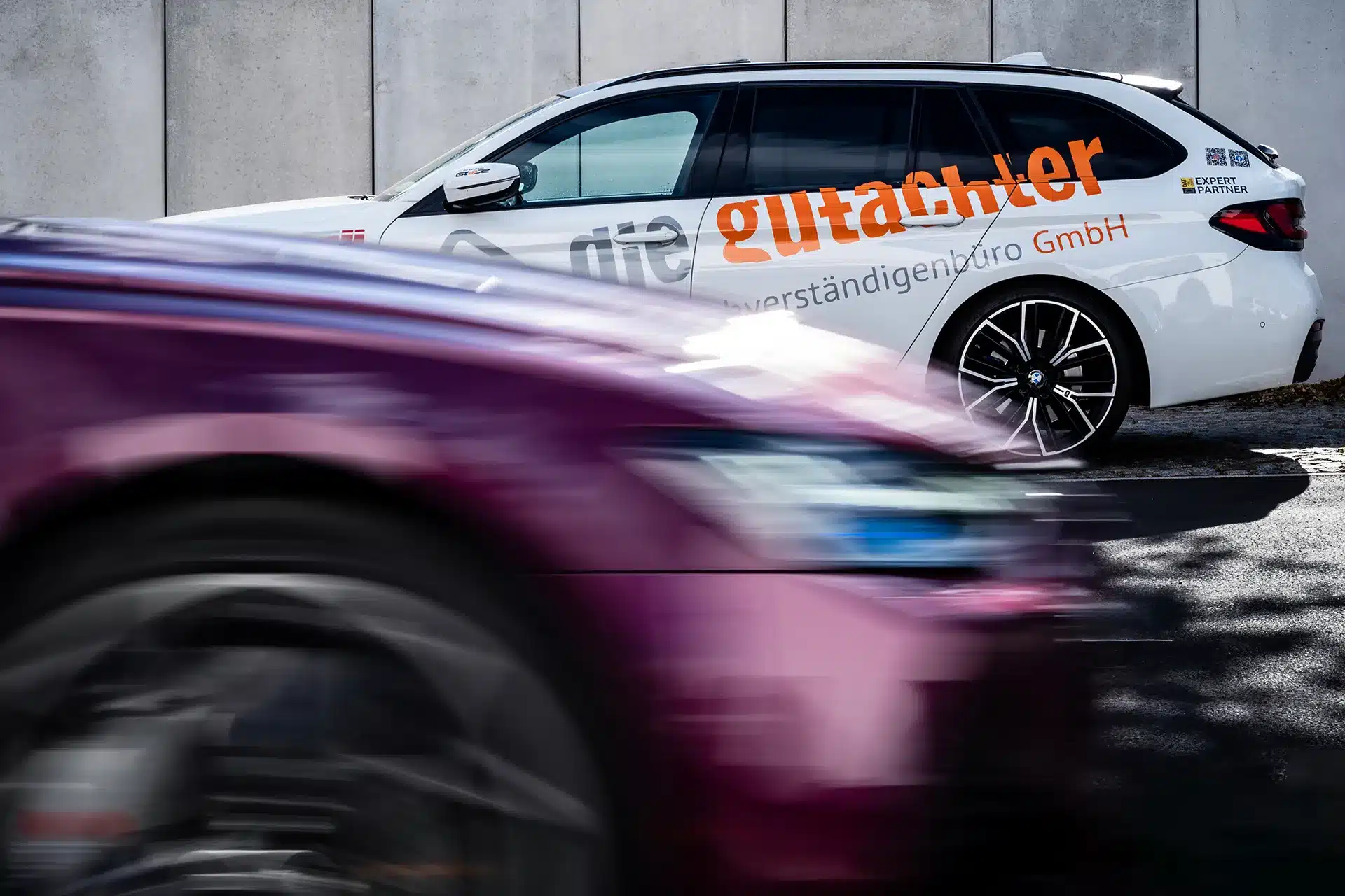 Ein unscharfes kastanienbraunes Auto fährt an einem sonnigen Tag vor einem geparkten weißen Kombi mit großer orangefarbener und grauer Schrift an der Seite in der Nähe einer Betonmauer vorbei.