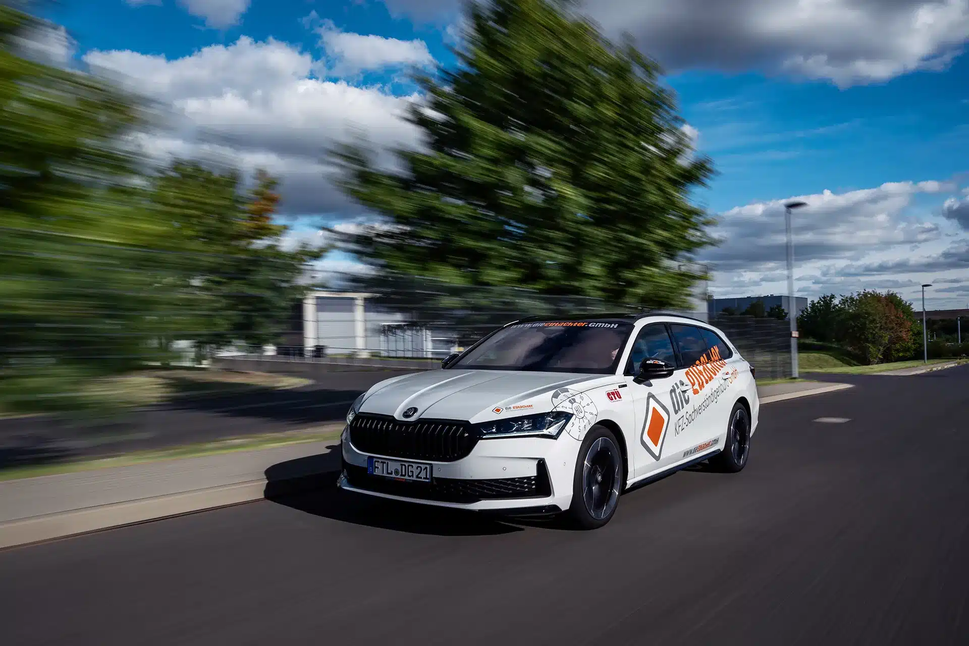 Ein weißes Škoda-Auto mit "Drive Score"-Schriftzug fährt schnell auf einer Straße, mit einem unscharfen Hintergrund aus Bäumen und Gebäuden unter einem teilweise bewölkten Himmel.