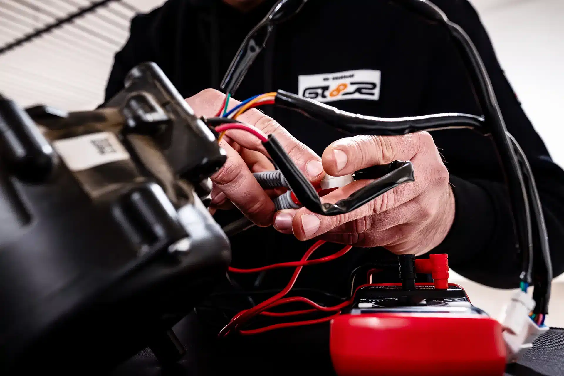 Eine Person prüft mit einem Multimeter elektrische Drähte, die mit einem schwarzen Gerät verbunden sind. Der Fokus liegt auf ihren Händen und dem Gerät, im Vordergrund sind Drähte und Werkzeuge zu sehen.