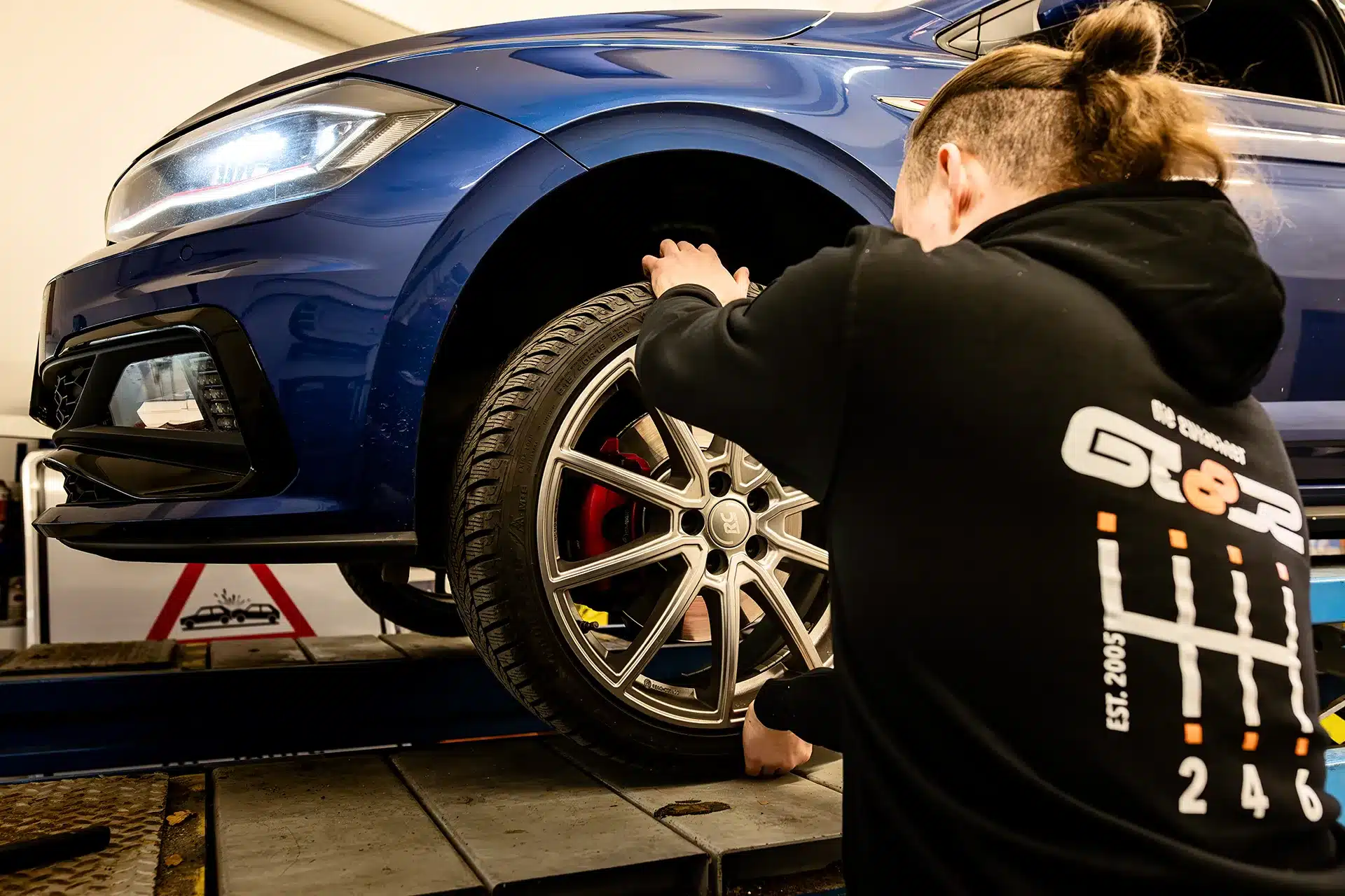 Eine Person sichert das Vorderrad eines blauen Autos in einer Garage, wahrscheinlich bei Wartungs- oder Reparaturarbeiten, während sie einen schwarzen Kapuzenpullover mit einer Grafik auf dem Rücken trägt.
