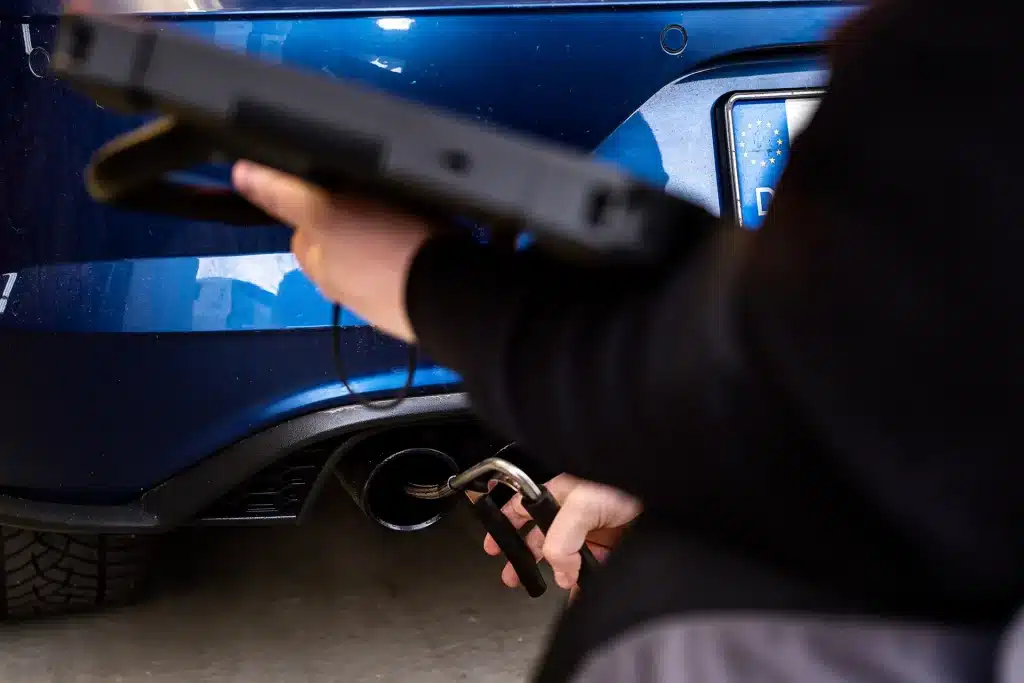 Eine Person hält ein Gerät in der Nähe des Auspuffrohrs eines blauen Autos und verwendet eine Ausrüstung zur Messung von Emissionen oder zur Durchführung von Diagnosen. Die Heckpartie und das Nummernschild des Fahrzeugs sind teilweise sichtbar.