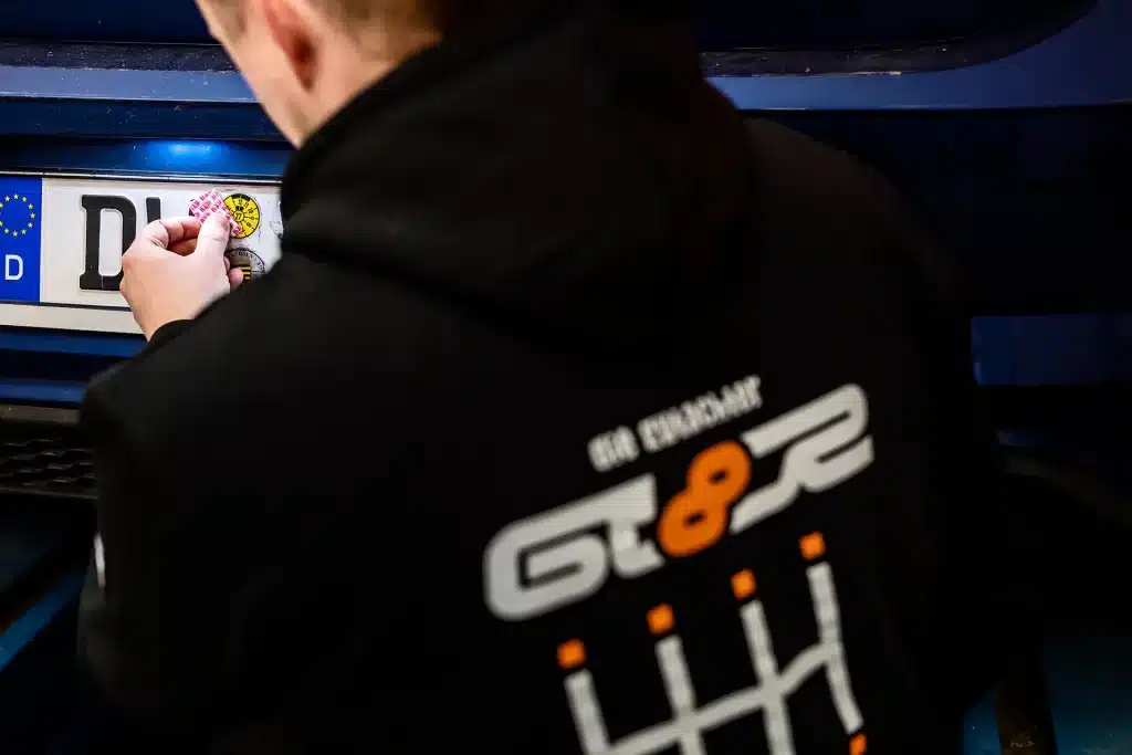 Eine Person in einem schwarzen Kapuzenpullover mit Schalthebelgrafik klebt einen runden Aufkleber auf das Nummernschild eines Autos. Der Schwerpunkt liegt auf der Anbringung des Aufklebers, wobei das Auto und die Kleidung deutlich zu sehen sind.