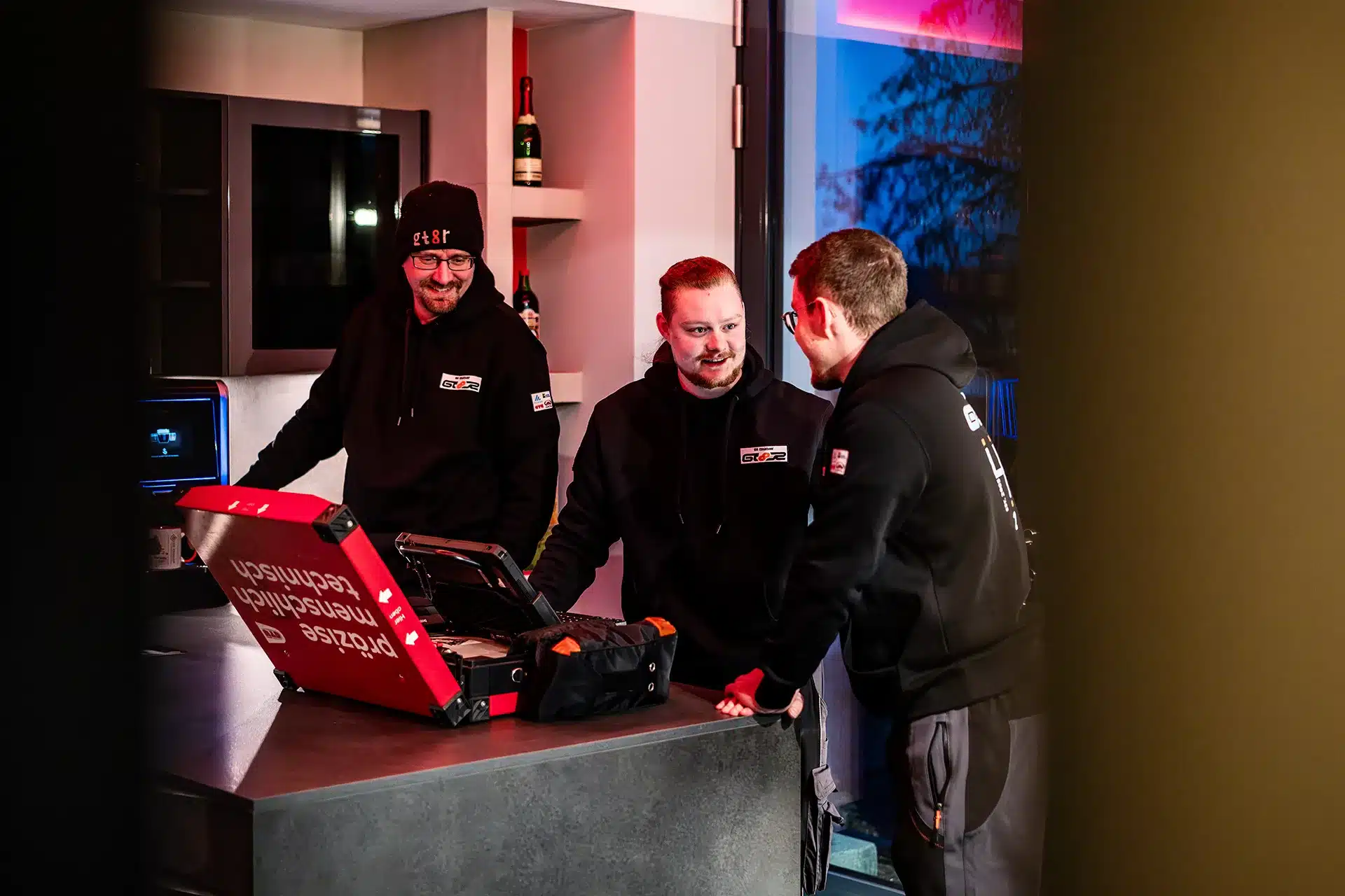 Drei Männer in schwarzen Kapuzenpullovern stehen in einer modernen Küche um einen Tresen herum, unterhalten sich und schauen auf einen offenen roten Gerätekoffer. Die Szene ist warm beleuchtet, mit Flaschen und Küchenutensilien im Hintergrund.