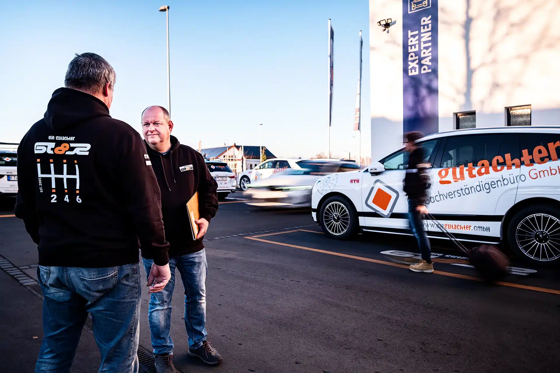 Zwei Männer stehen auf einem Parkplatz in der Nähe eines weißen Lieferwagens mit orangefarbener Aufschrift und unterhalten sich, während eine Person mit einem Koffer vorbeigeht. Die Szene scheint sich vor einem Geschäft oder einer Fachwerkstatt abzuspielen.