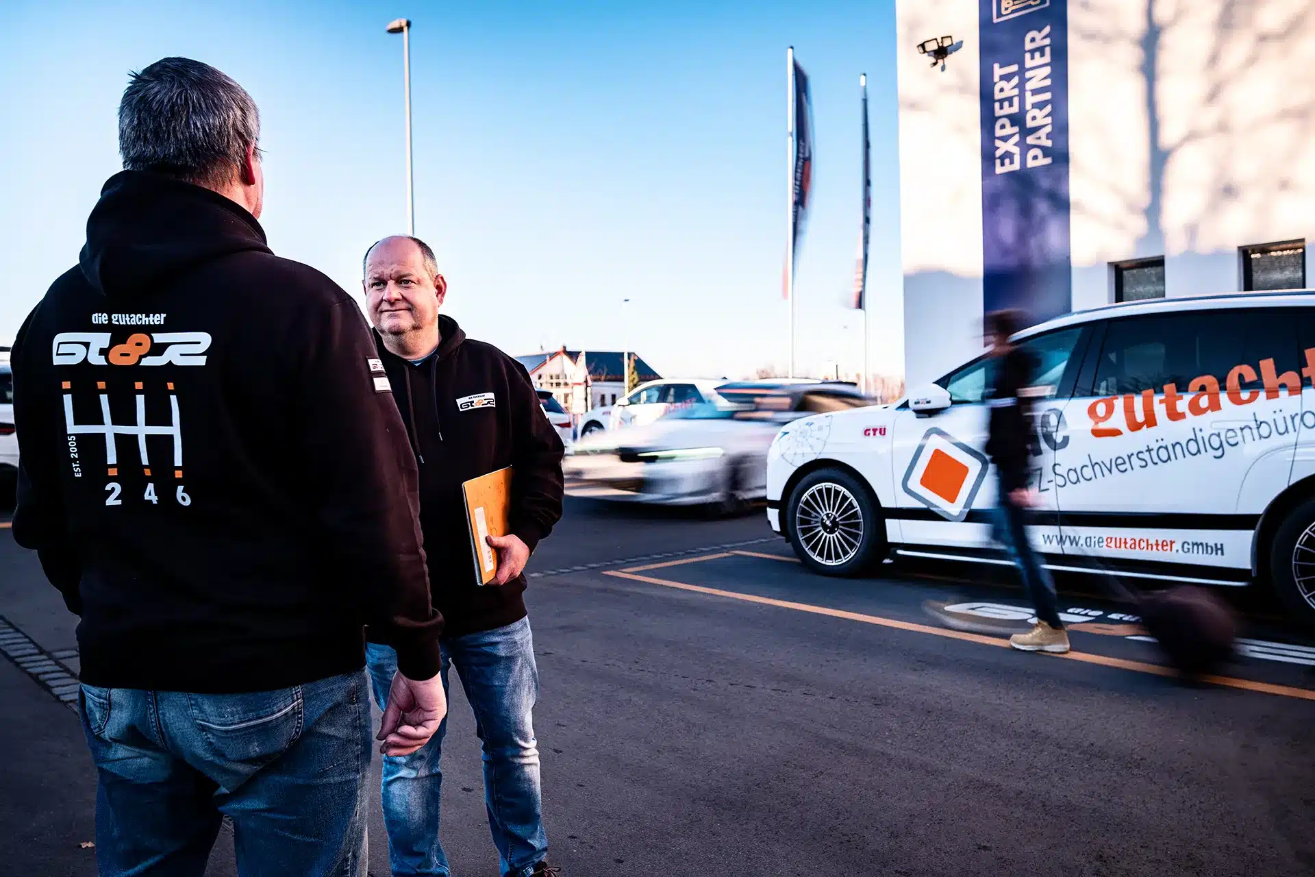 Zwei Männer stehen auf einem Parkplatz neben einem weißen Auto mit orangefarbenen Logos und deutschem Text und unterhalten sich. Ein Mann hält einen Ordner. Im Hintergrund sind ein Gebäude, Fahnen und unscharfe Fußgänger zu sehen, die vorbeigehen.