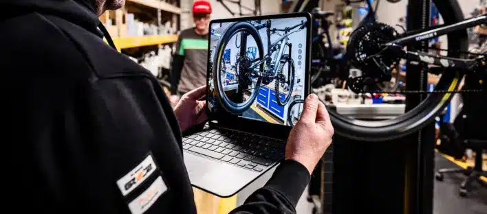 Eine Person hält einen Laptop in der Hand, auf dem ein Fahrradrad abgebildet ist. Sie steht in einer Werkstatt, in der Fahrradwerkzeuge und -teile zu sehen sind, während eine andere Person im Hintergrund arbeitet.