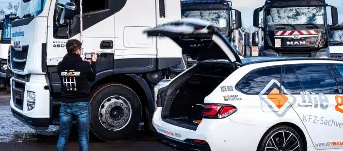 Eine Person steht neben einem weißen Inspektionsfahrzeug mit geöffnetem Kofferraum und fotografiert einen großen Lastwagen, der zwischen mehreren anderen Lastwagen auf einem Industriegelände geparkt ist. Die Fahrzeuge sind mit Firmenlogos und Schriftzügen versehen.