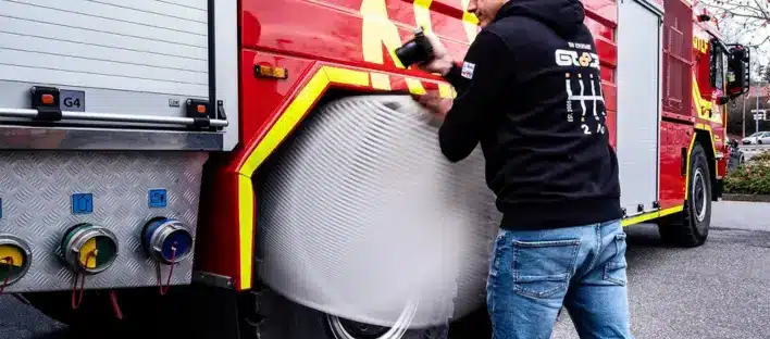 Eine Person in schwarzem Kapuzenpulli und Jeans reinigt die Seite eines roten Feuerwehrautos und macht dabei kreisende Bewegungen mit einem Reinigungswerkzeug in der Nähe des Rades. Das Feuerwehrauto hat sichtbare Schläuche und Metallplatten.