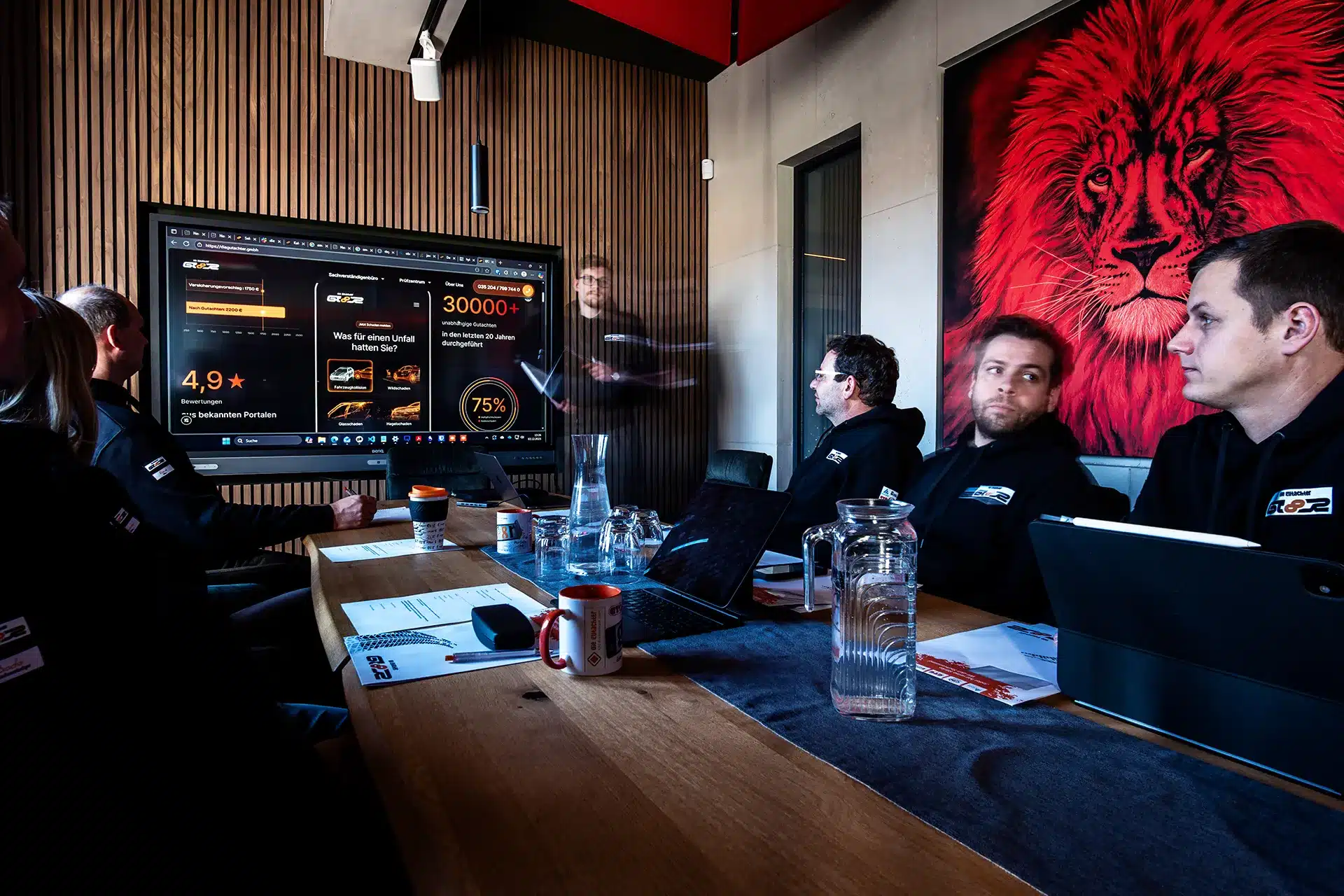 Sechs Personen sitzen in einem modernen Besprechungsraum mit Laptops und Papieren um einen Tisch und sehen sich eine Präsentation auf einem großen Bildschirm an. Ein roter Löwe hängt an der Wand hinter ihnen. Eine Person steht und wirkt leicht verschwommen.