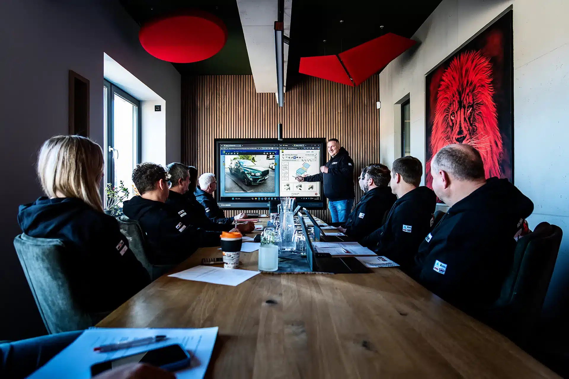 Eine Gruppe von Menschen in schwarzen Kapuzenpullovern sitzt um einen Konferenztisch und beobachtet aufmerksam, wie ein Moderator auf einem großen Bildschirm in einem modernen Konferenzraum Bilder von Autos erklärt.