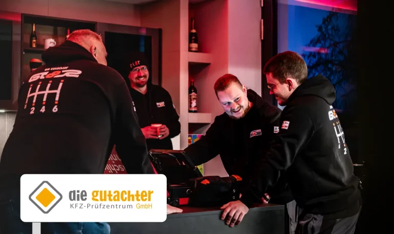 Vier M&auml;nner in schwarzen Kapuzenpullovern stehen l&auml;chelnd um einen Tresen und unterhalten sich in einem modernen Raum mit Theke und Regalen. In der Ecke steht das Logo der "die Gutachter GT8R KFZ-Pr&uuml;fzentrum GmbH". In der Mitte der KFZ Pr&uuml;fer erkennt man einen Koffer mit Werkzeug.