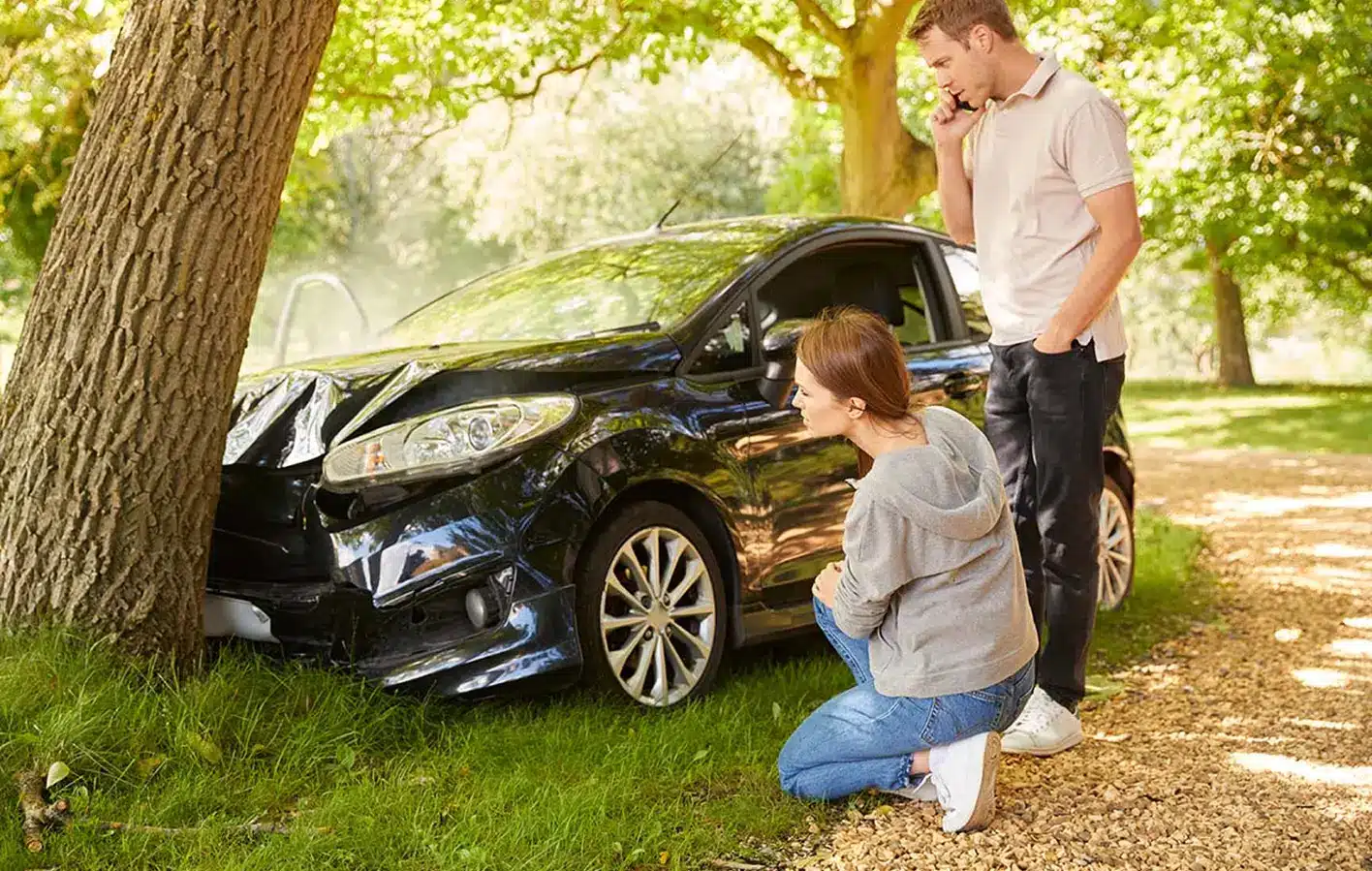 Ein Mann telefoniert mit seinem Handy, während eine Frau neben einem schwarzen Auto kniet, das gegen einen Baum geprallt ist; beide wirken besorgt und fragen sich wahrscheinlich: Was tun nach Unfall? Die Szene spielt in einem sonnigen, bewaldeten Gebiet.